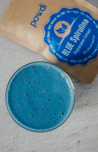 Blue spirulina smoothie in a glass with blue spirulina powder packet on a light surface